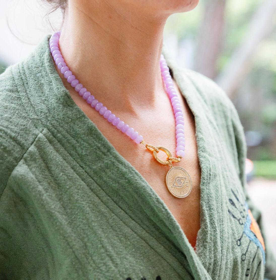 A girl wearing a light pink stone beaded necklace with a gold chain link in the middle with a gold engraved eye medallion attached to it