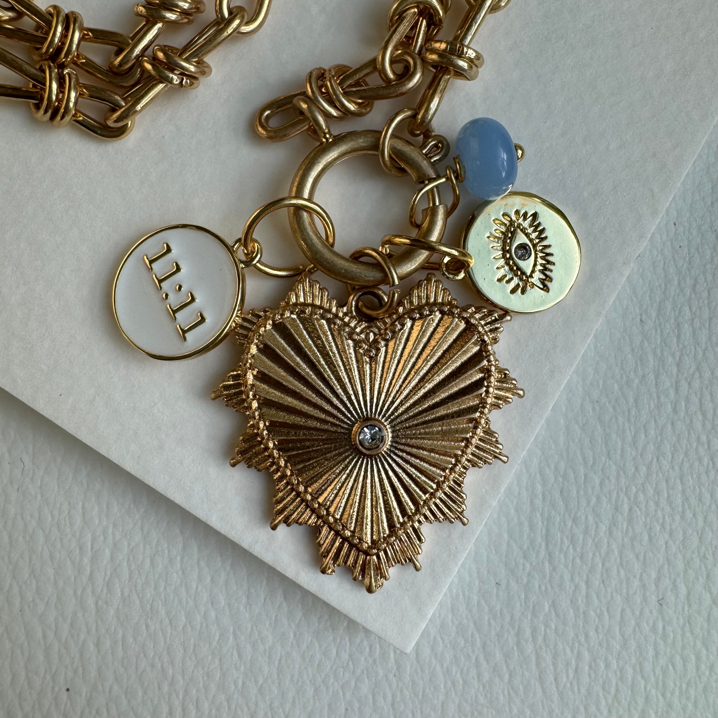 Picture of gold chain link charm necklace. Necklace has a large gold heart pendant with a diamond at its center. Three smaller charms include a blue bead, a round white charm with a gold eye and a white charm reading, "11:11".