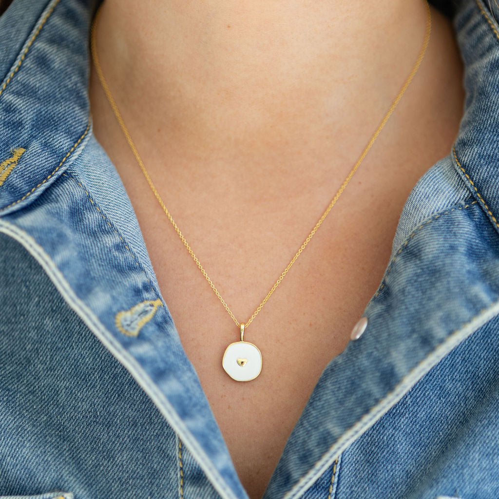 Gold necklace with a round white enamel pendant worn by a person in a denim jacket.