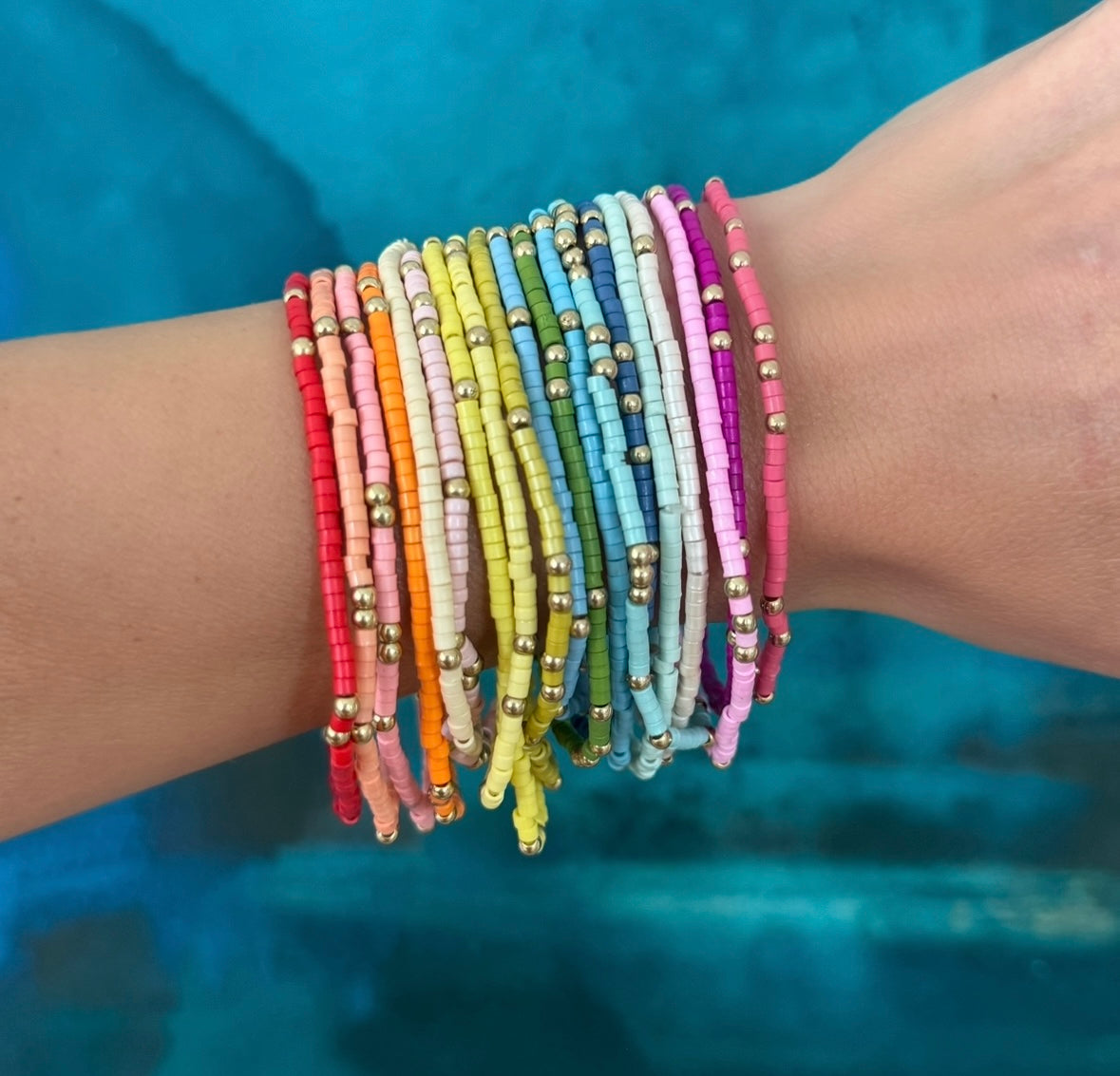 Picture of a wrist wearing 19 different colored seed bead bracelets with accent gold beads. Bracelets are red, orange, beige, pinks, white, yellows, blues, greens and purple.
