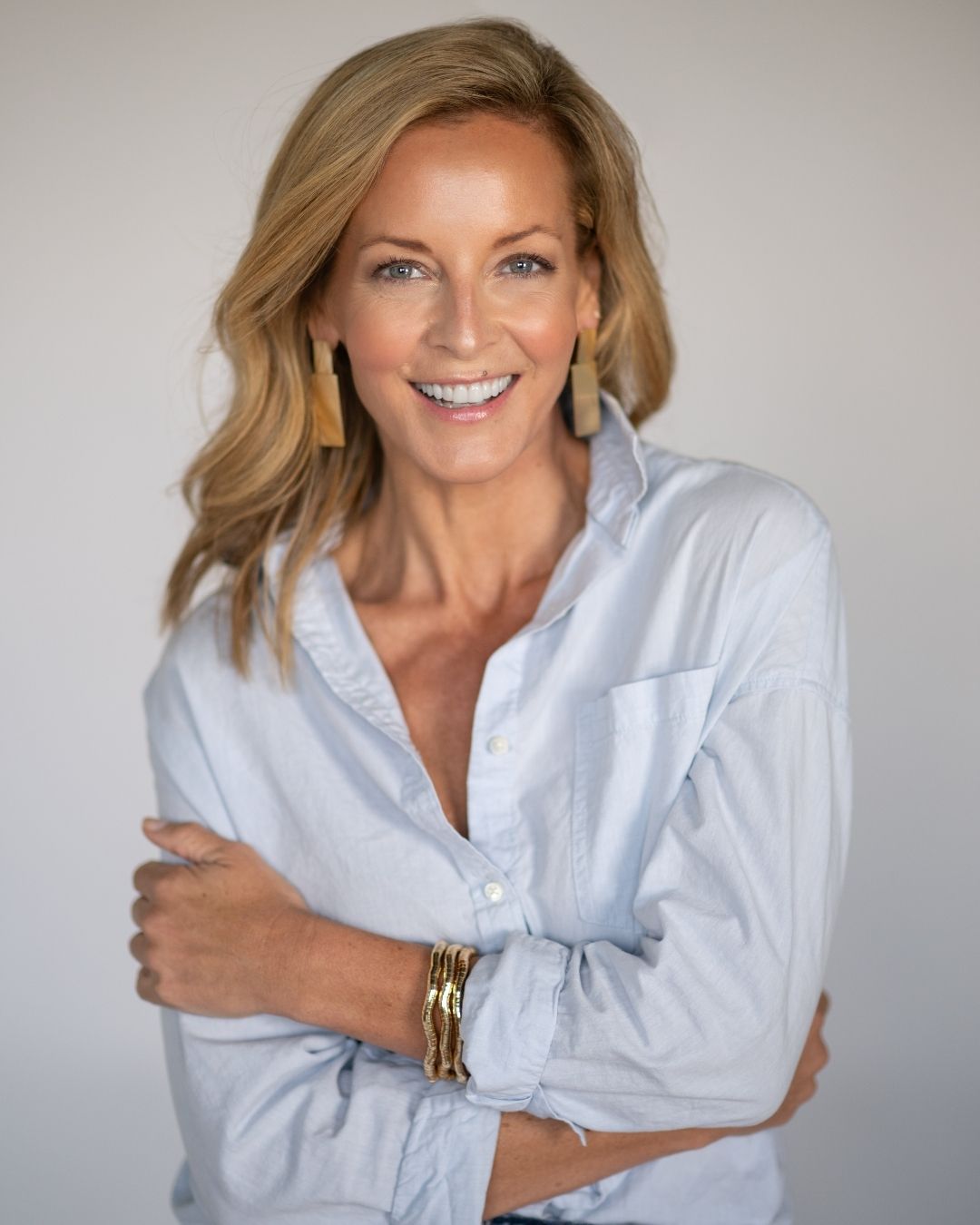 Woman wearing a light blue button up shirt with large gold statement earrings and bracelets on a plain background