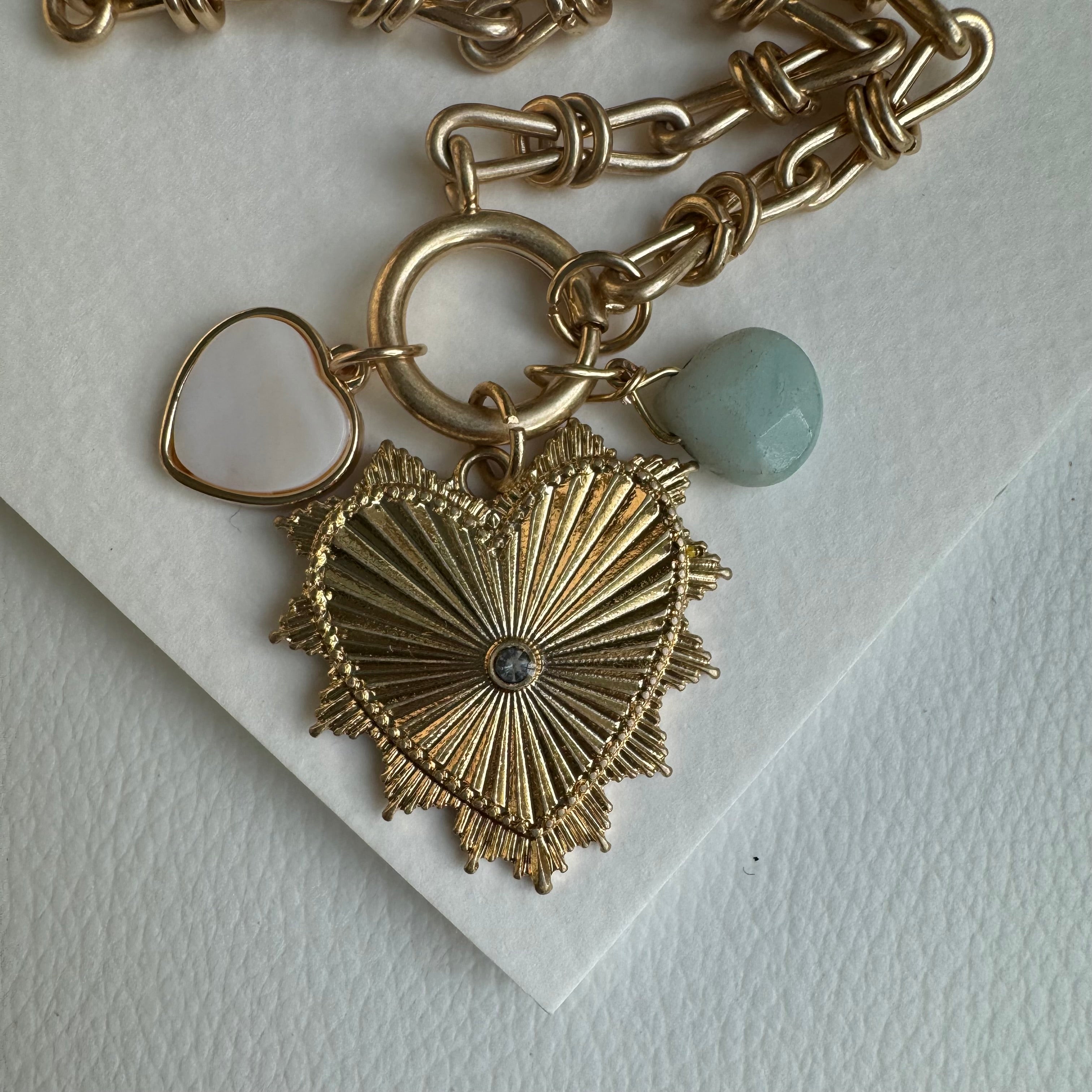 Picture of gold chain link charm necklace. Necklace has a large gold heart pendant with a diamond at its center. Two smaller charms include a muted turquoise bead and a white heart charm with a gold border.