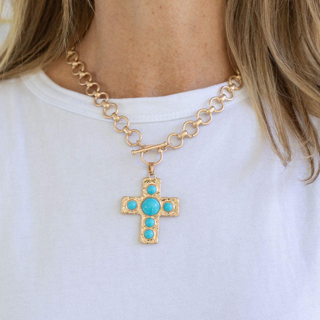 Picture of a woman wearing a circular chain link necklace with a toggle clasp. Necklace has a large gold Cross with turquoise beads inlaid in it.