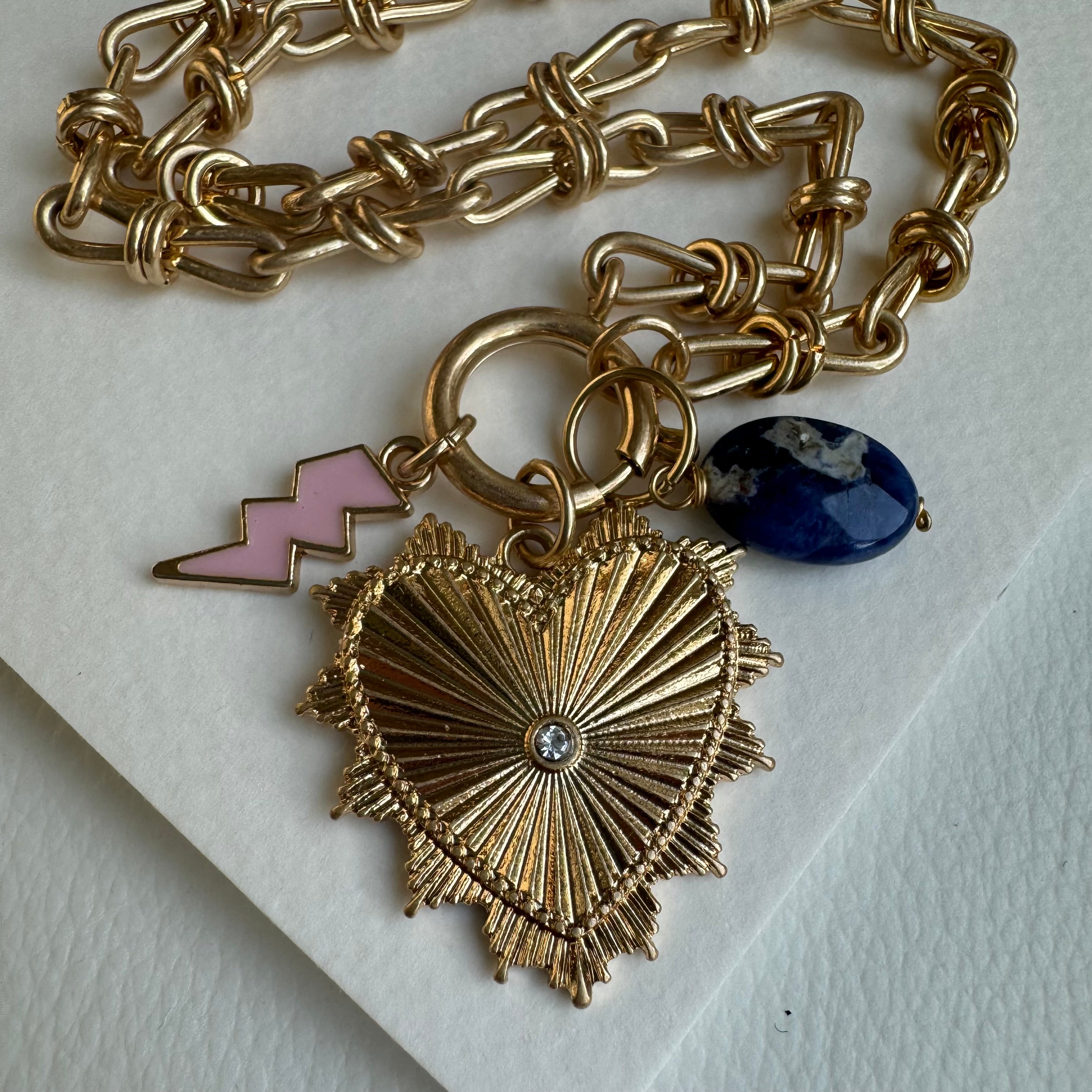 Picture of gold chain link charm necklace. Necklace has a large gold heart pendant with a diamond at its center. Two smaller charms include a dark blue bead and a pink lightening bolt.