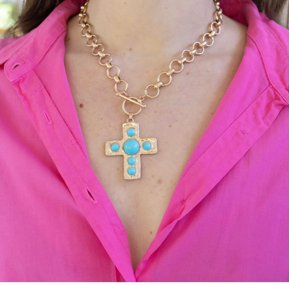 Large round link gold necklace with a toggle clasp and a cross pendant featuring turquoise stones worn by a person in a pink shirt.