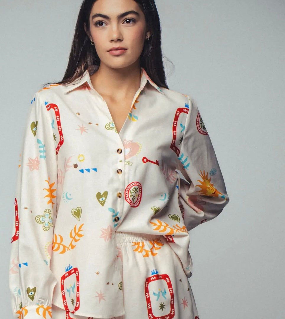 Picture of a woman wearing a white silk button up top with matching pants. Clothing set has bright shapes and plant and sun themed pictures.
