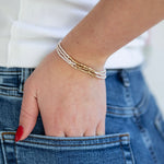 Hand wearing a white and gold bead bracelet set with blue jeans and a white shirt in the background
