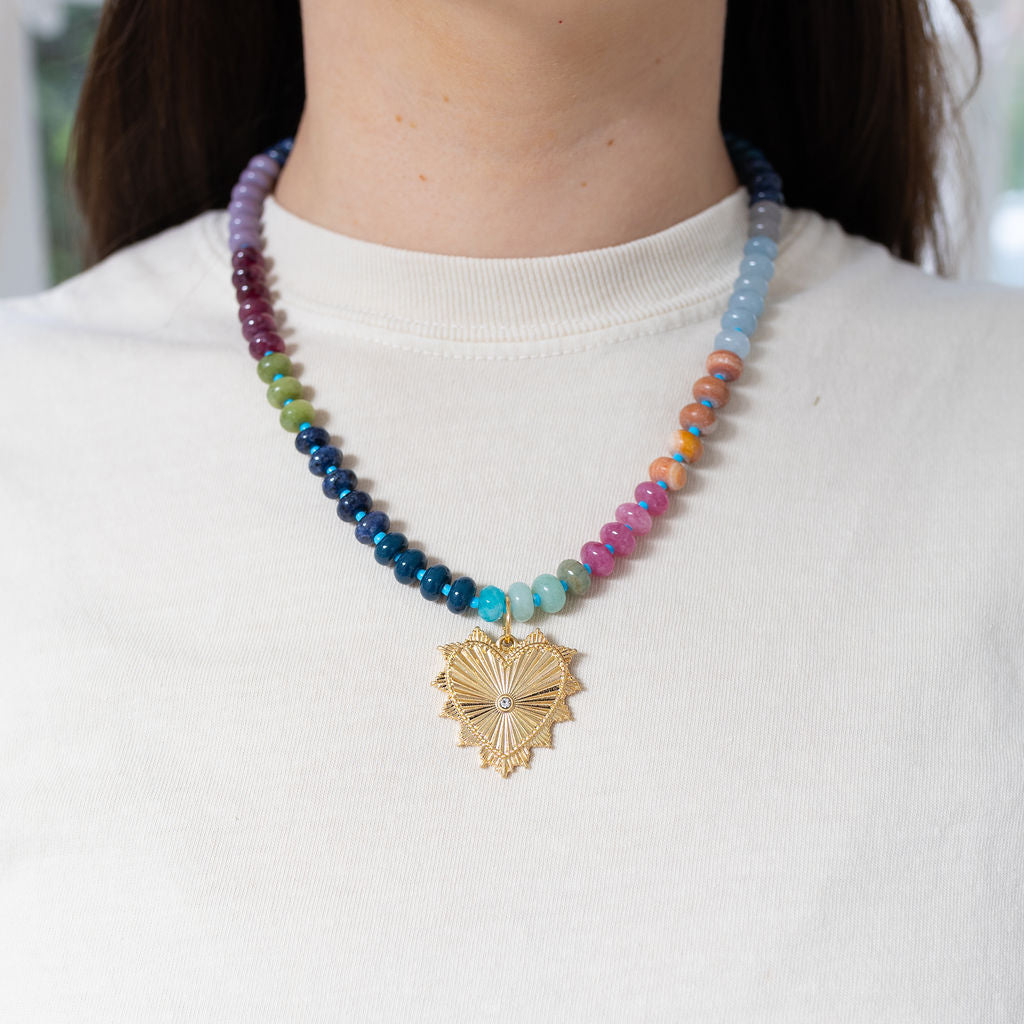 Picture of model wearing a multi-colored beaded necklace of blues, pink, orange, green and purples. Necklace also has large gold heart pendant with a diamond in its center.