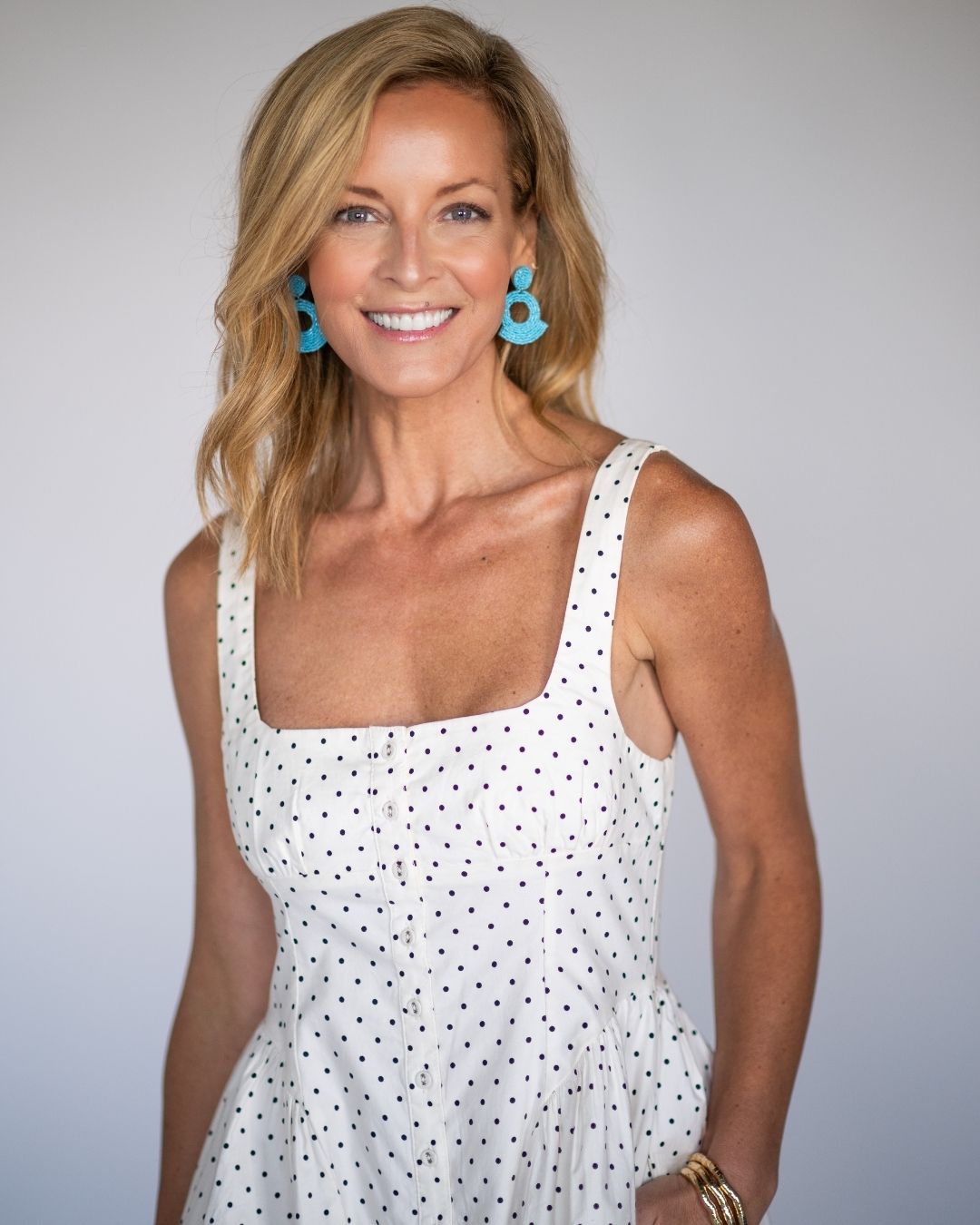 Woman wearing a white polka dot dress with blue statement earrings against a plain background