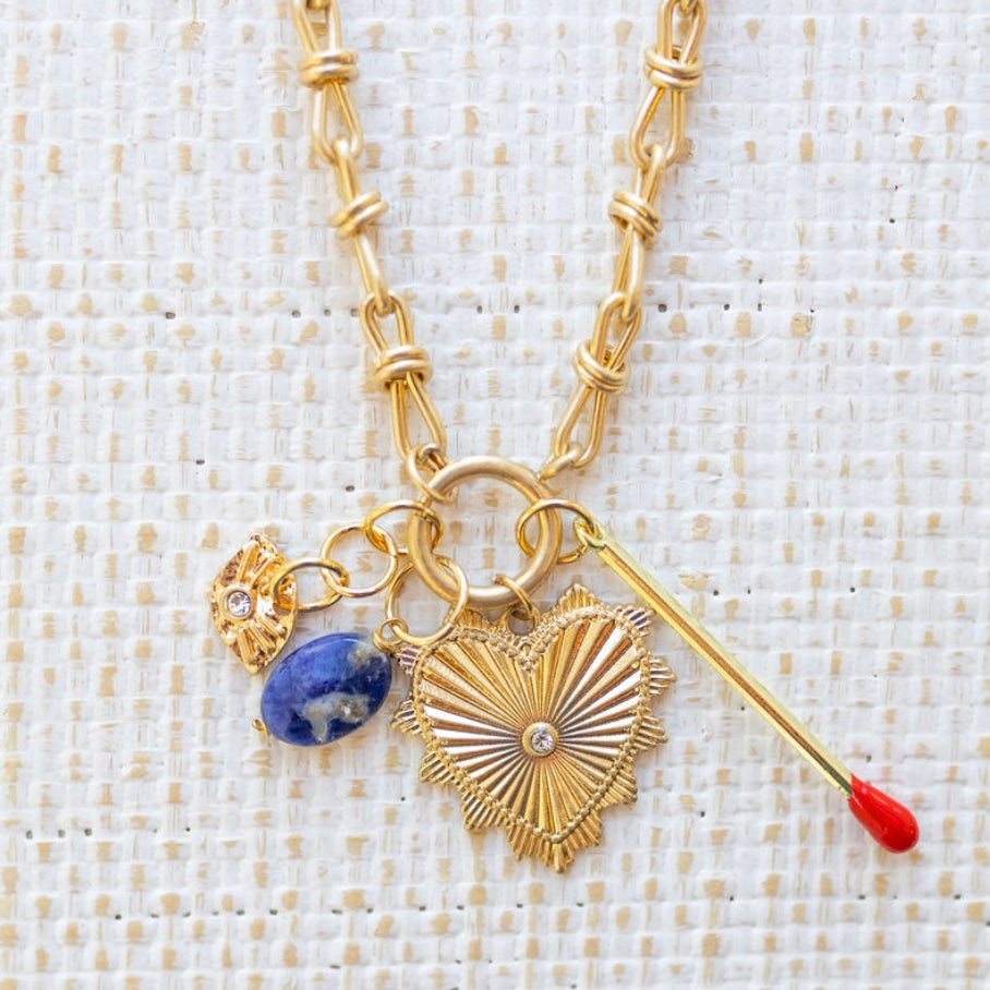 Picture of gold toggle chain with a large gold heart pendant with a diamond at the center. Necklace has three additional charms, a gold and diamond evil eye, a blue marbled bead and a gold matchstick.