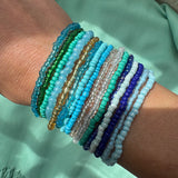 Multiple beaded bracelets in blues and greens on a wrist with a blurred green background