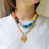 Model wearing large multicolored beaded necklace stacked with front clasping gold chain necklace with a heart pendant