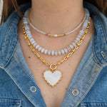 A three layered necklace set include a tiny beaded necklace in white, a larger resin beaded necklace in light blue and a gold chain with a large white heart pendant
