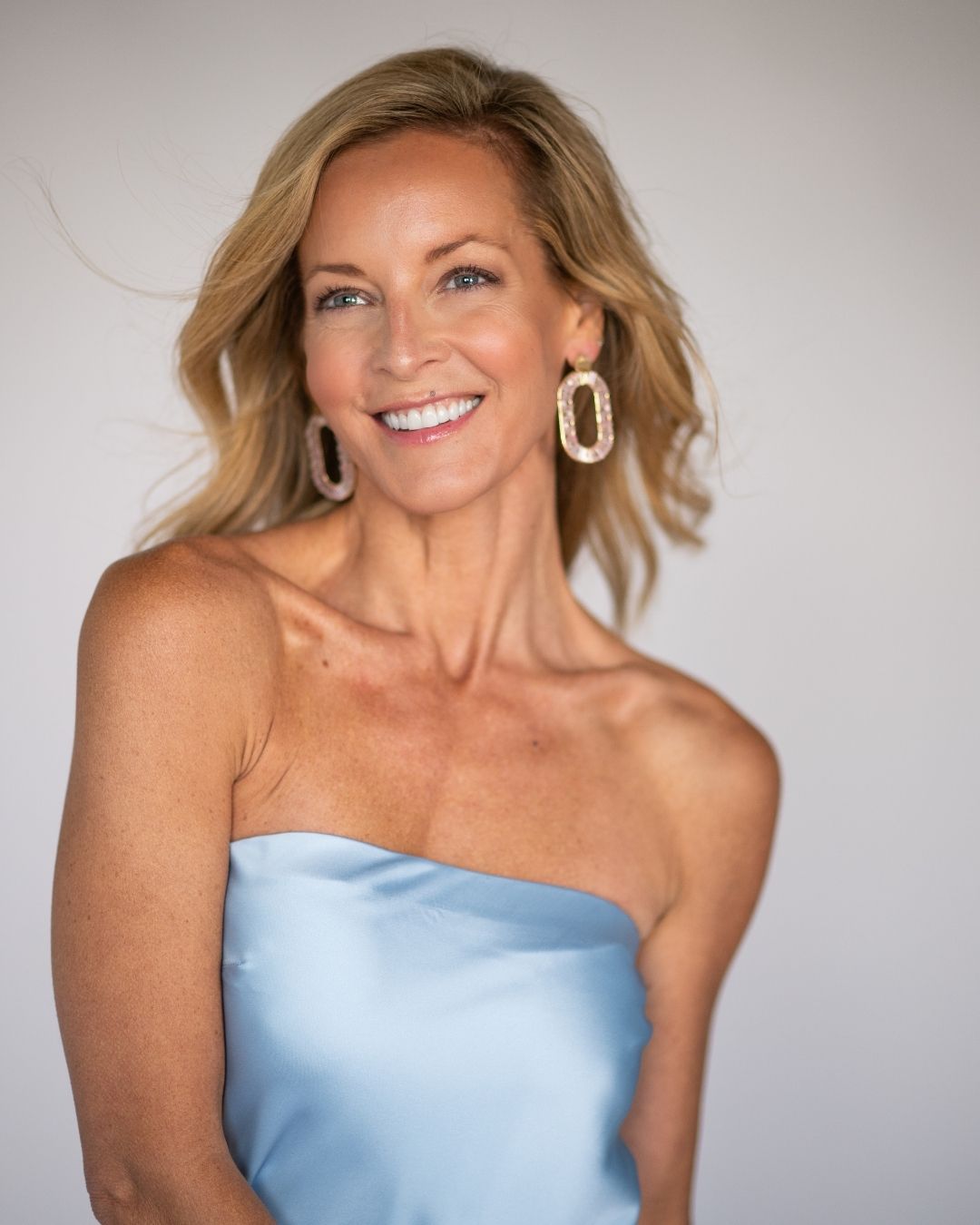 Woman wearing a strapless light blue dress with large gold hoop earrings against a plain background