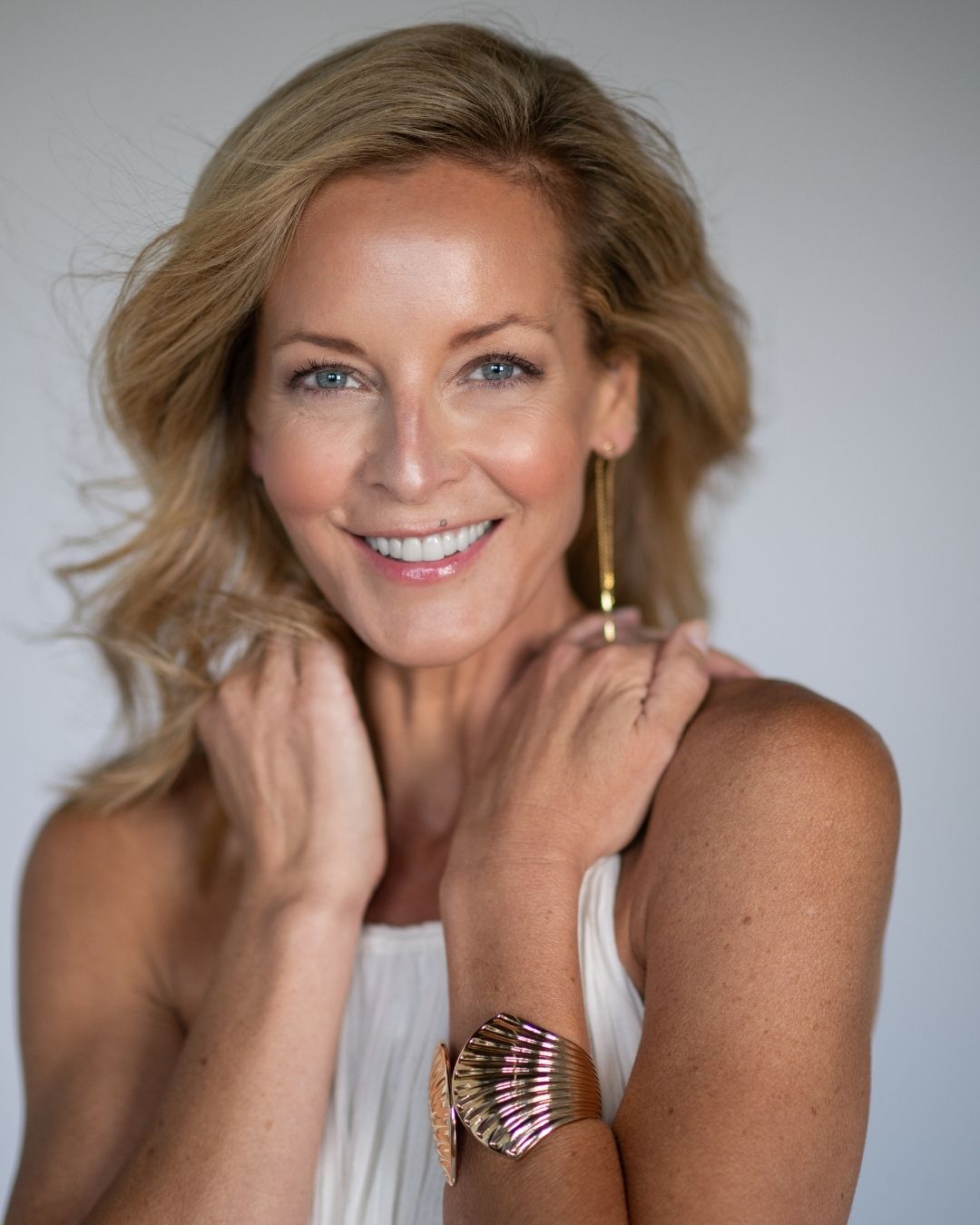 Woman with blonde hair and a white top, wearing a gold clam shell cuff bracelet and gold dangling earrings, against a plain background