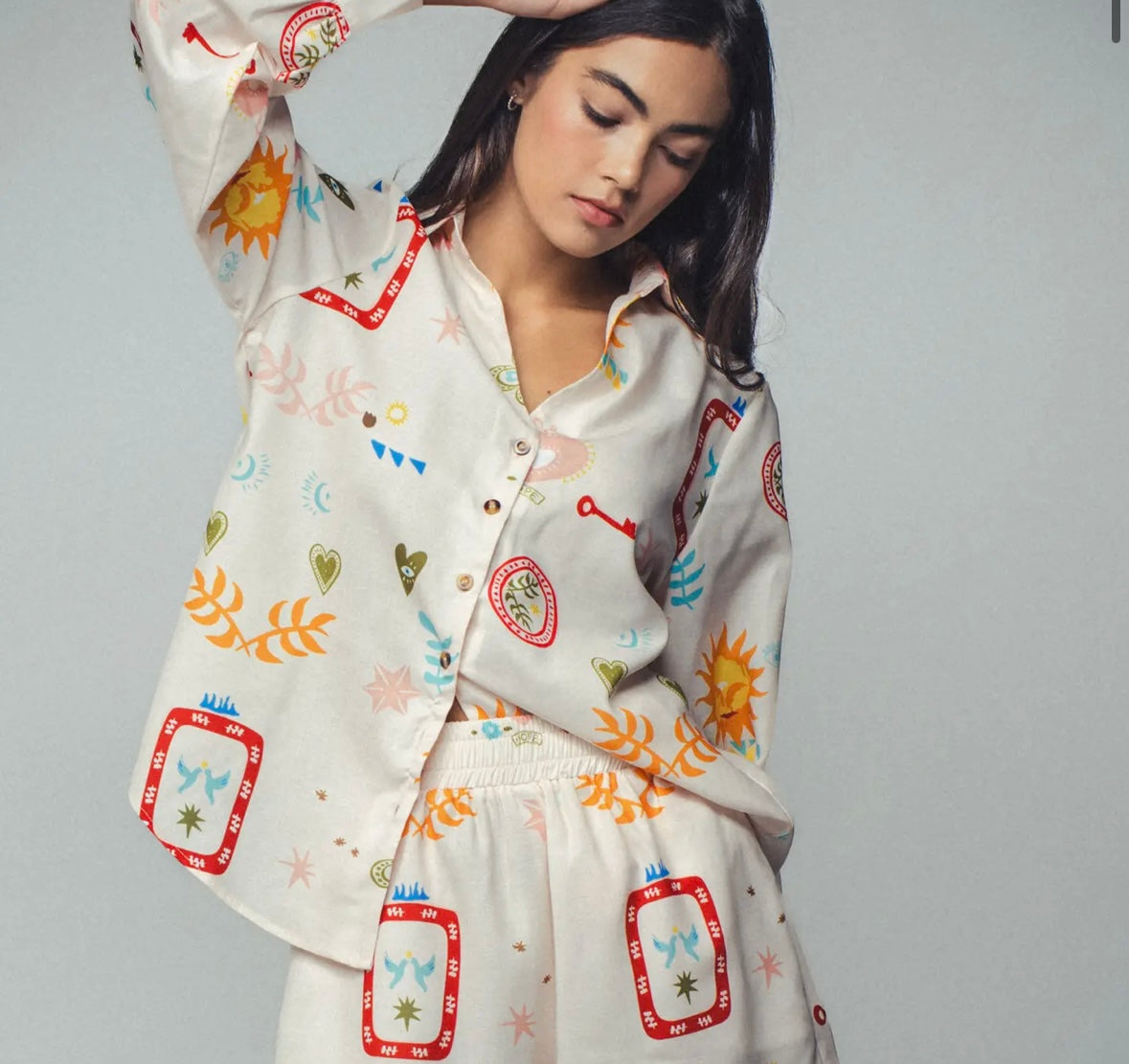 Picture of a woman wearing a white silk button up top with matching pants. Clothing set has bright shapes and plant and sun themed pictures.
