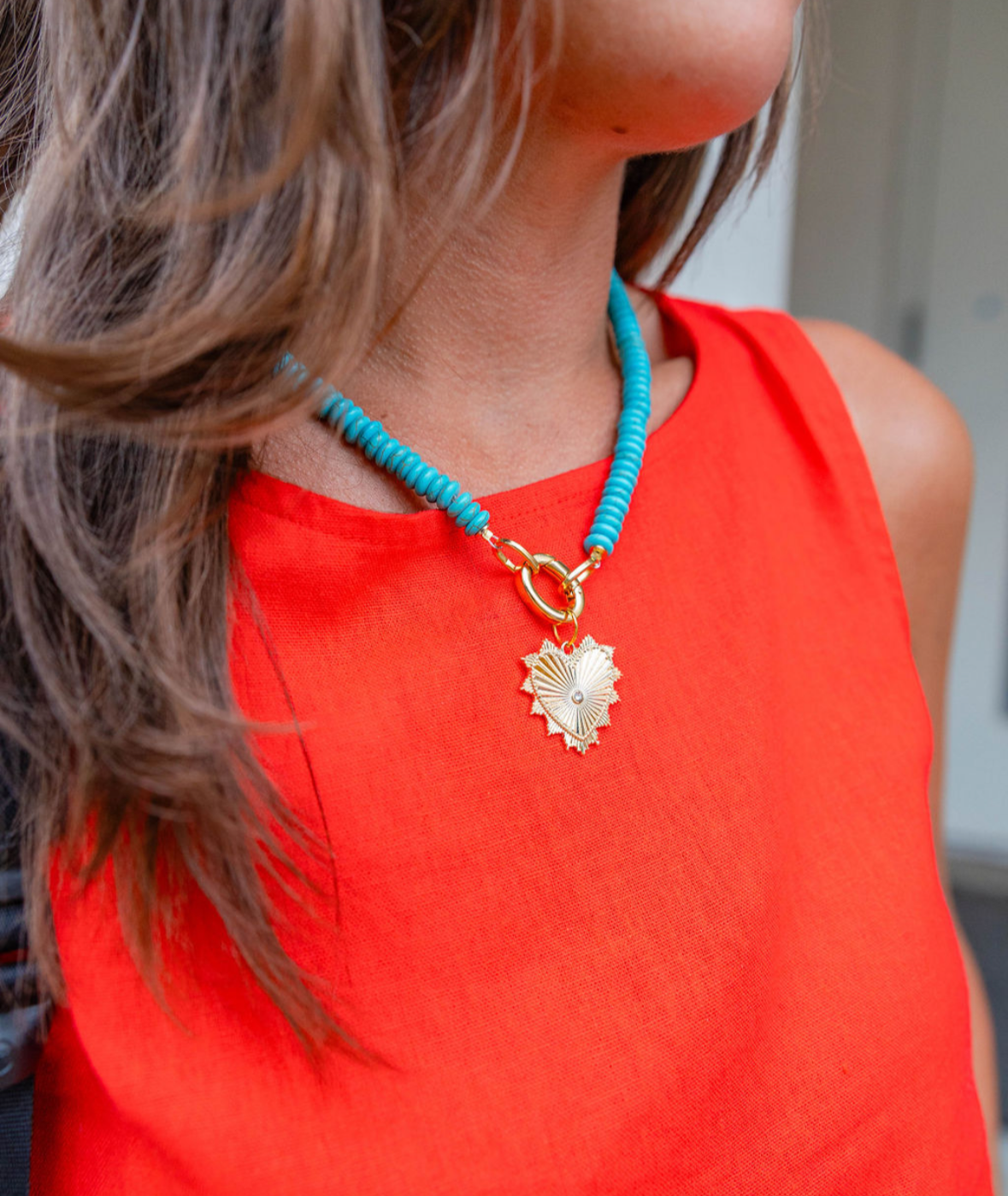 Woman wearing a big blue beaded necklace with a gold heart shape medallion attached to it