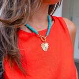 Woman wearing a big blue beaded necklace with a gold heart shape medallion attached to it