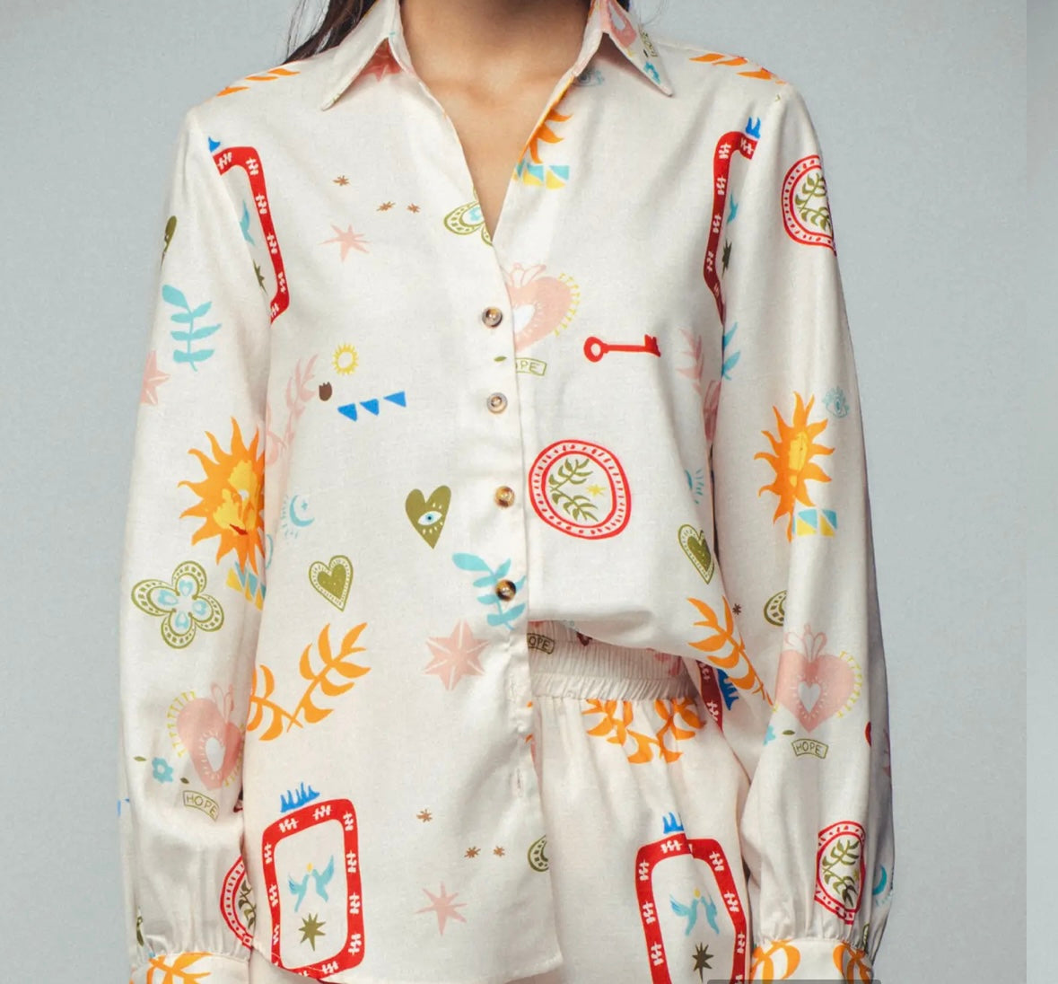 Picture of a woman wearing a white silk button up top with matching pants. Clothing set has bright shapes and plant and sun themed pictures.