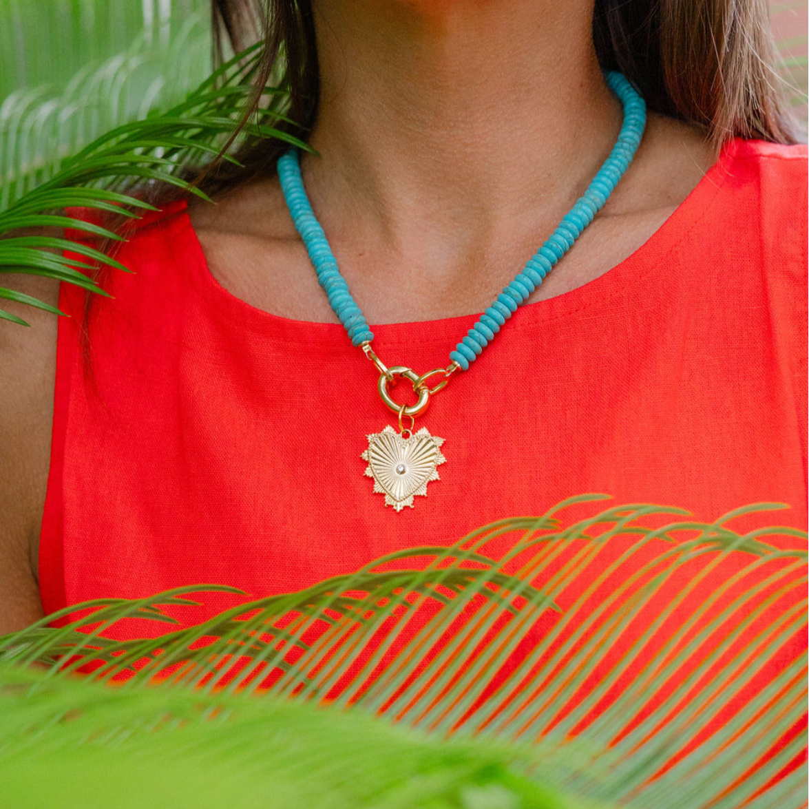 Woman wearing a big blue beaded necklace with a gold heart shape medallion attached to it