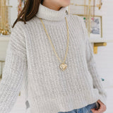 Woman in a white turtleneck wearing a long gold chain with a gold Puffy heart pendant.