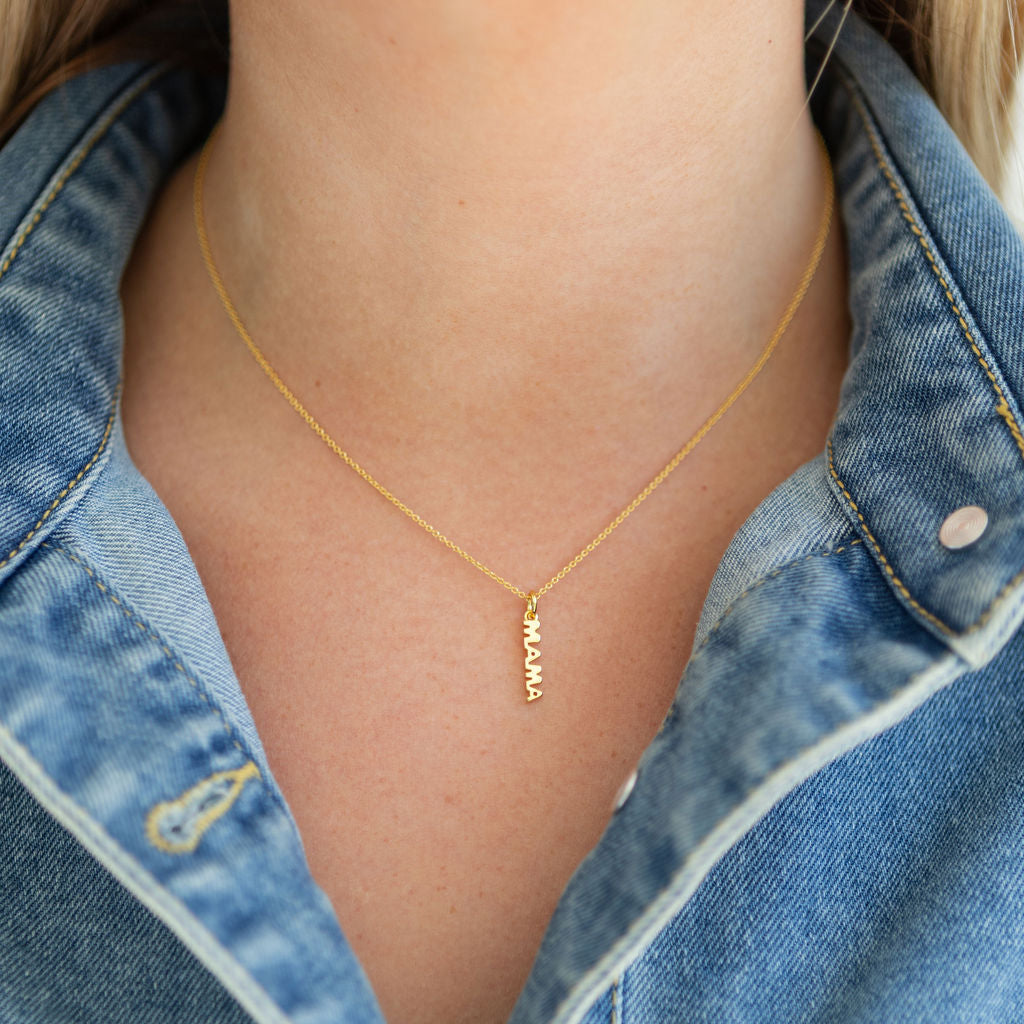 Delicate gold necklace with 'MAMA' pendant worn by a person in a denim jacket.