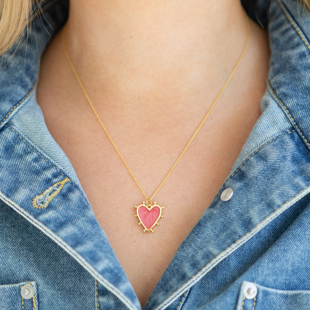 Gold necklace with a pink heart pendant worn by a person in a denim jacket.