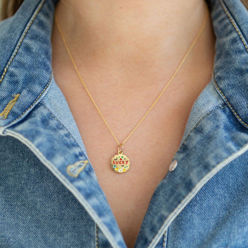 Gold necklace with a gold disc colorful pendant that reads 'Lucky' worn by a person in a denim shirt.