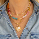 A person wearing three layered necklaces with colorful beads, a tiny beaded gold rectangle necklace and a larger gold link necklace with a toggle clasp and a gold-tone heart pendant.