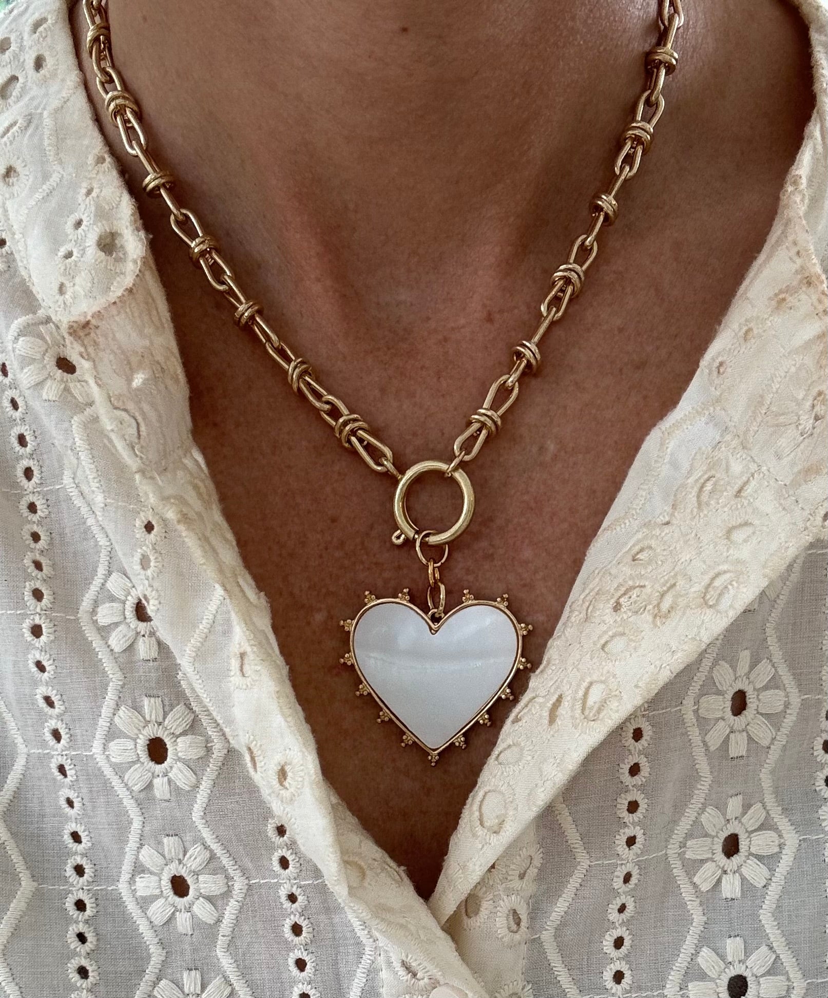 Model wearing a gold toggle chain with a large white heart pendant trimmed with gold studs.
