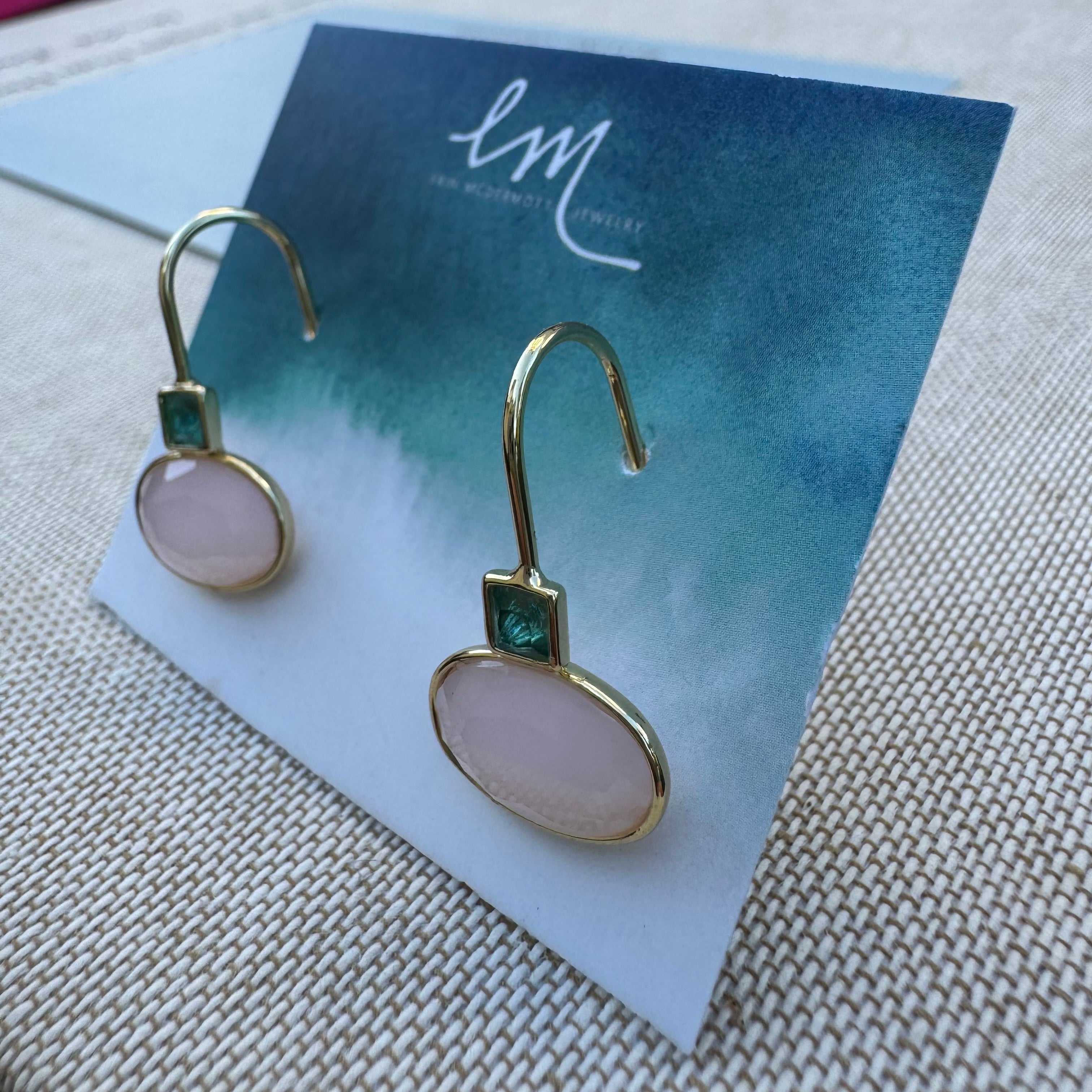 Gold dangle earrings with pink stones on a blue card with 'EM' branding.