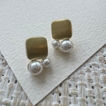 Gold square and pearl earrings on a textured white surface