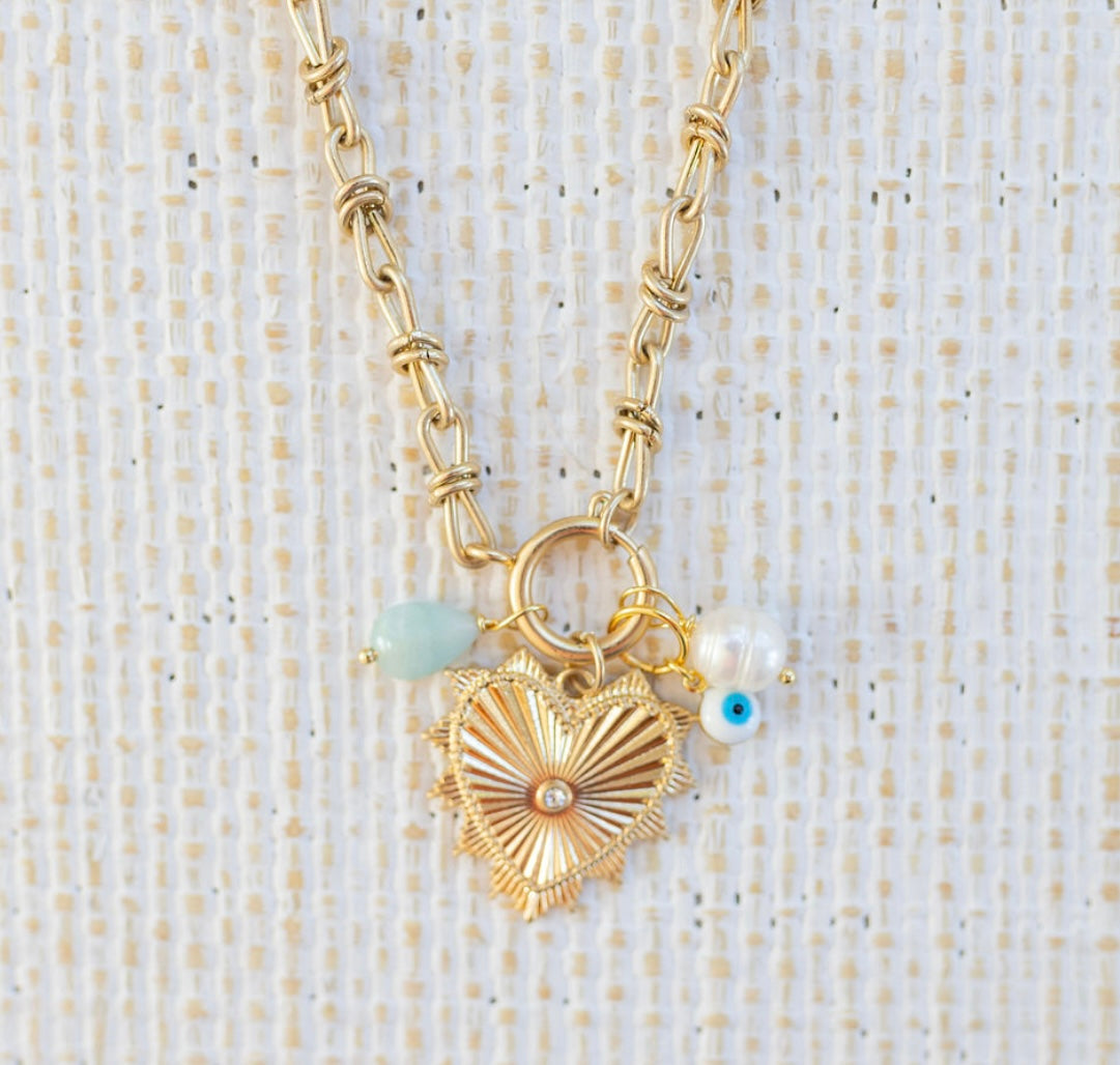Picture of gold toggle chain with large gold heart pendant with a diamond at the center. Necklace also has a light teal bead, a fresh water bead and an evil eye attached.