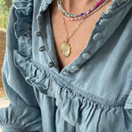 Picture of a woman wearing a blue blouse and four necklaces. 3 necklaces are dainty bead necklaces in pink and white, blue and light blue and white and gold. One necklace is a gold link chain necklace with a large textured gold disc pendant.
