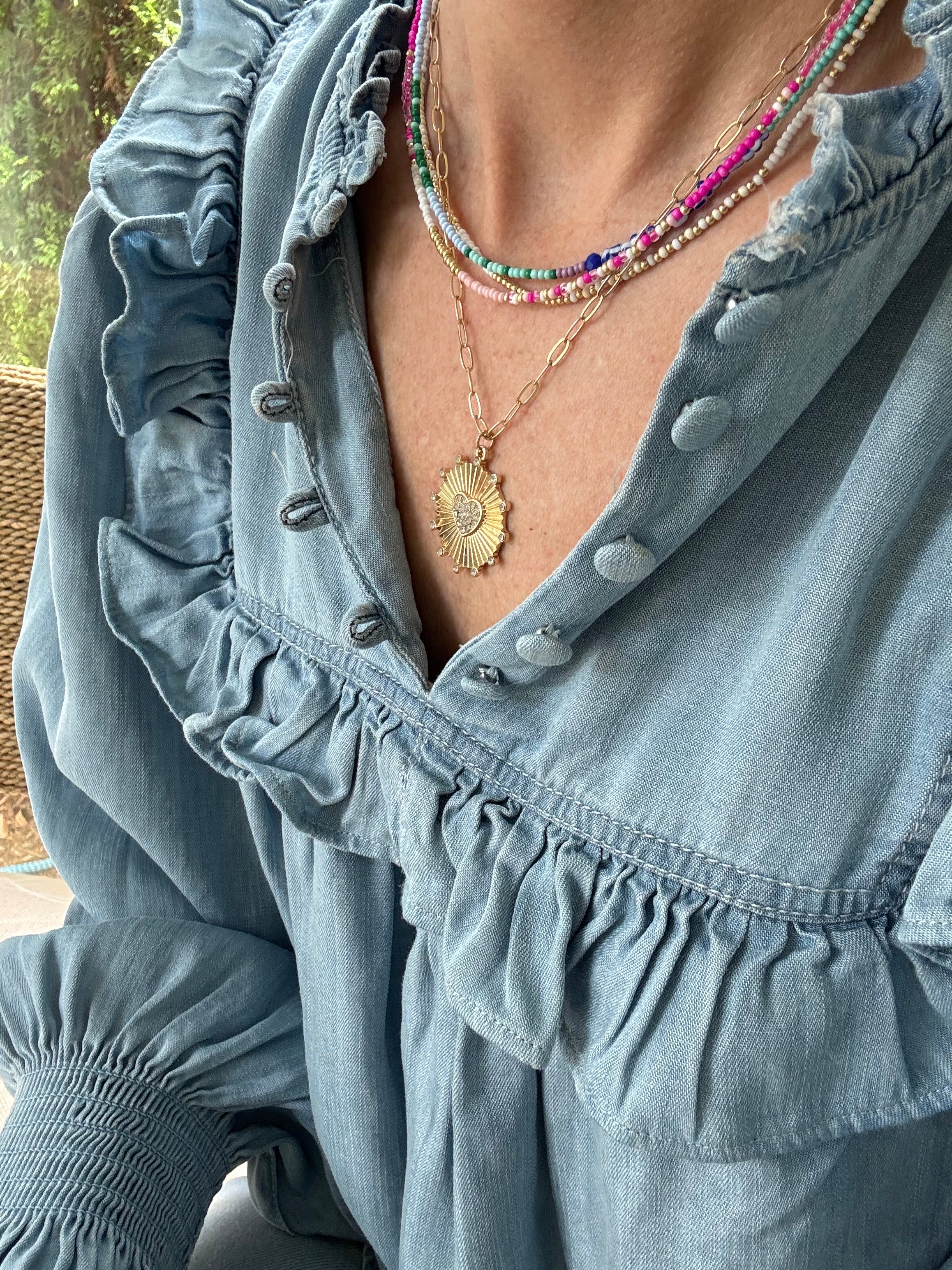 Picture of a woman wearing a blue blouse and four necklaces. 3 necklaces are dainty bead necklaces in pink and white, blue and light blue and white and gold. One necklace is a gold link chain necklace with a large textured gold disc pendant.