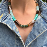 Necklace with multicolored turquoise, green and brown beads worn by a person in a denim jacket.