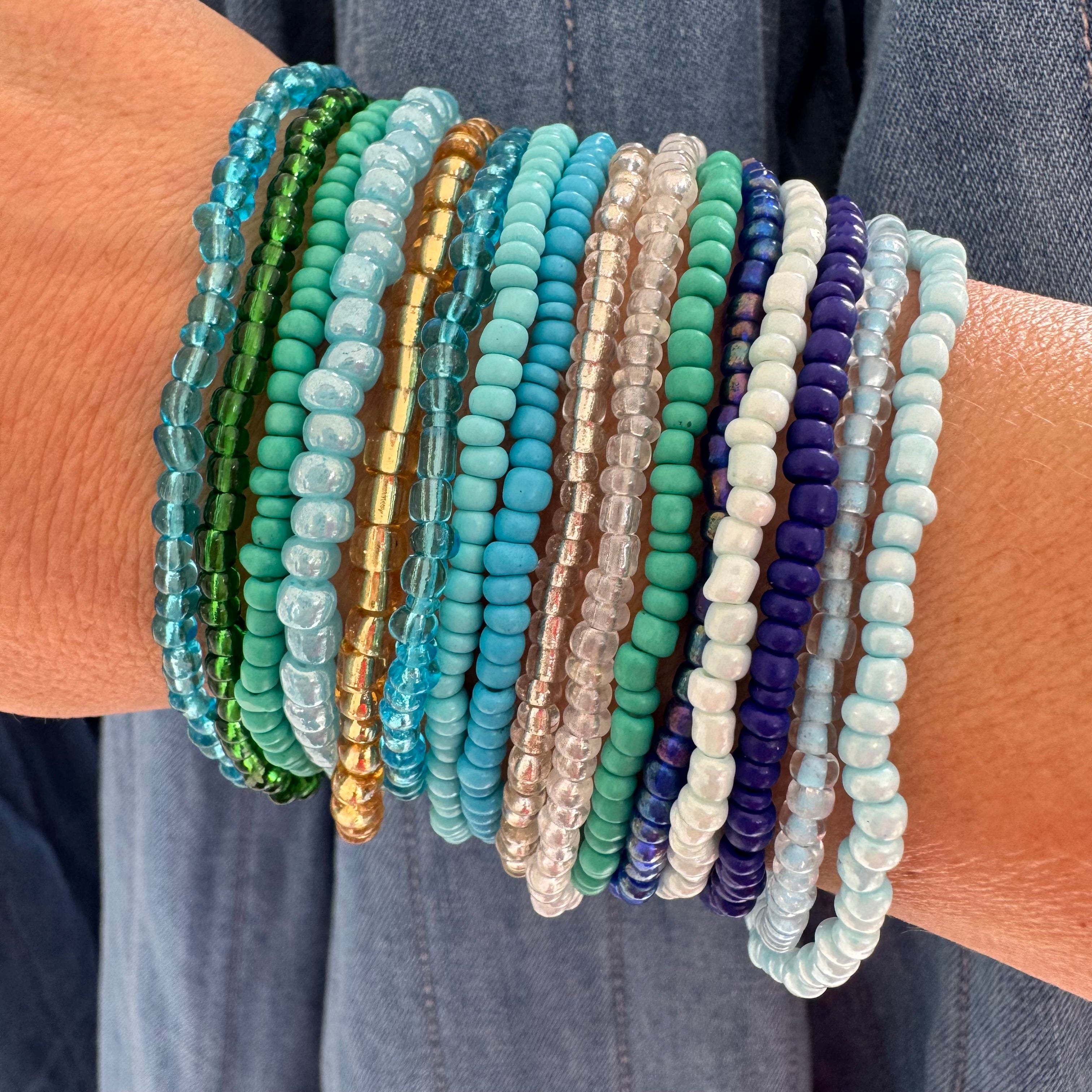 Multiple beaded bracelets of various blue, green and beige colors on a wrist against a denim background