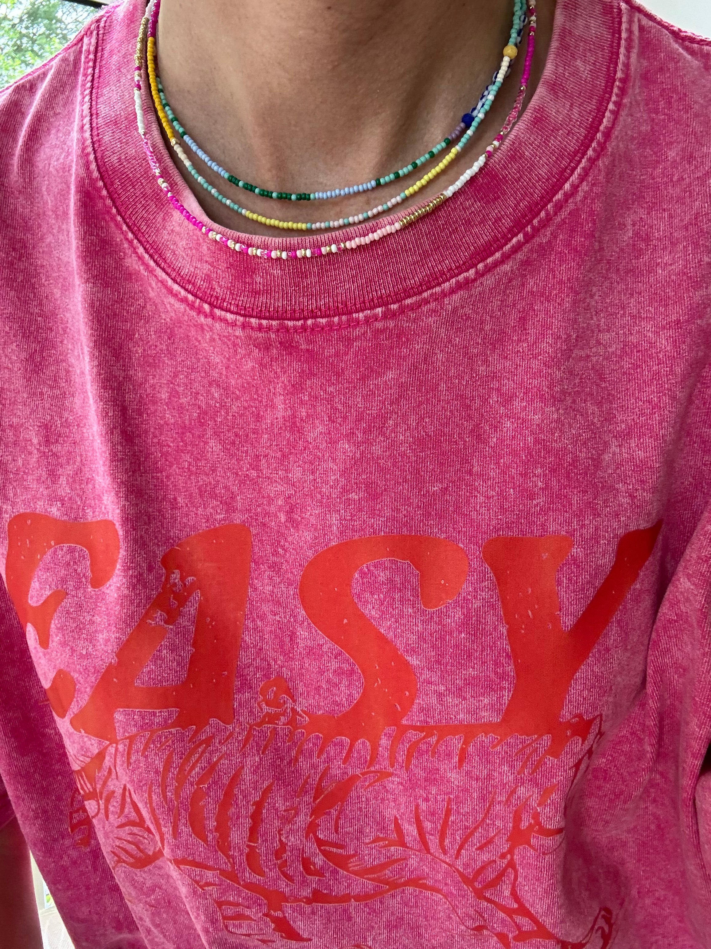 Picture of a woman wearing a pink Tshirt and three dainty bead necklaces. One necklace is yellow and white, one in dark and light blues and one in white and gold.