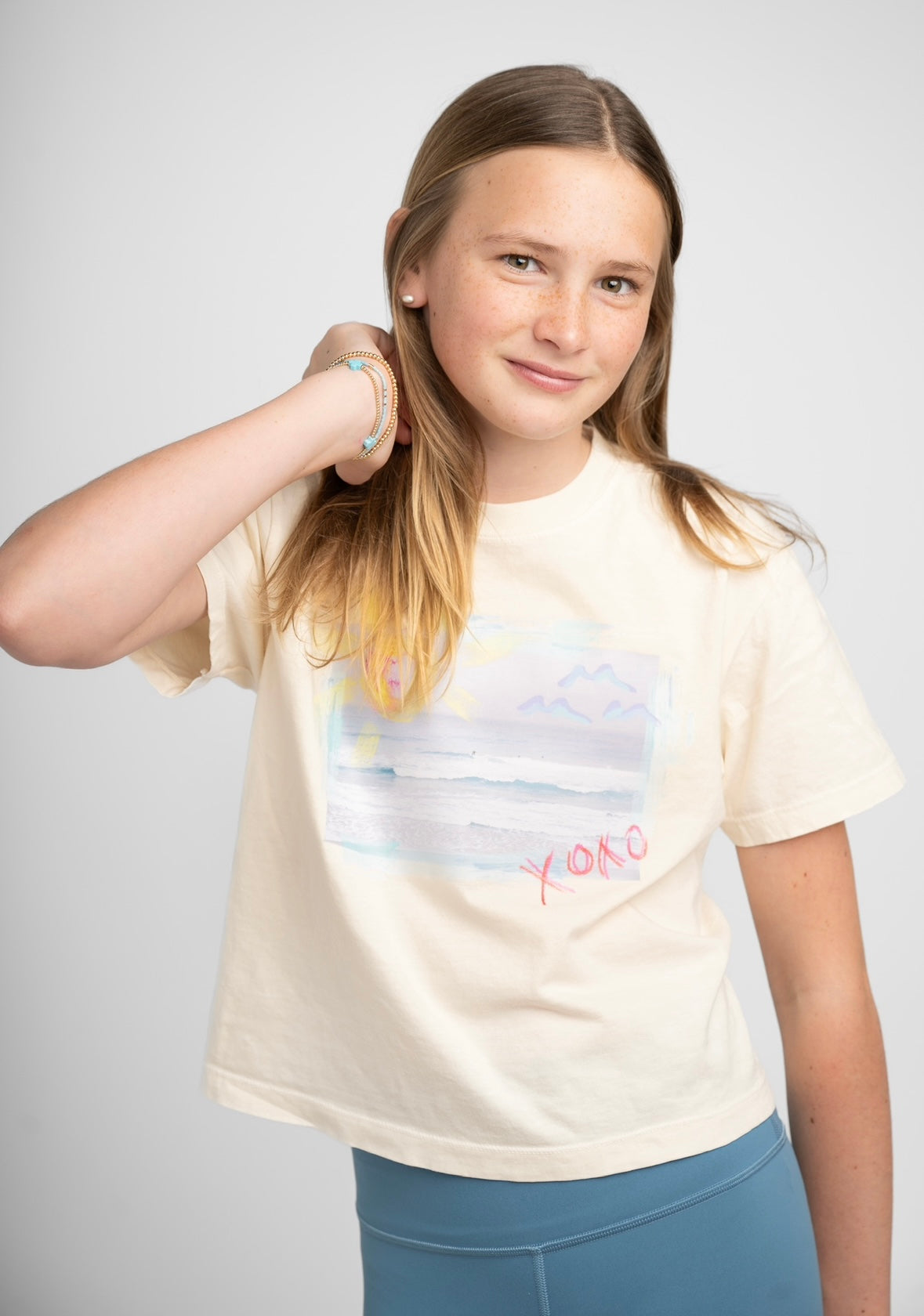 Picture of young girl wearing a cream colored Tshirt with a beach scene printed on the front with the letters XOXO in pink.