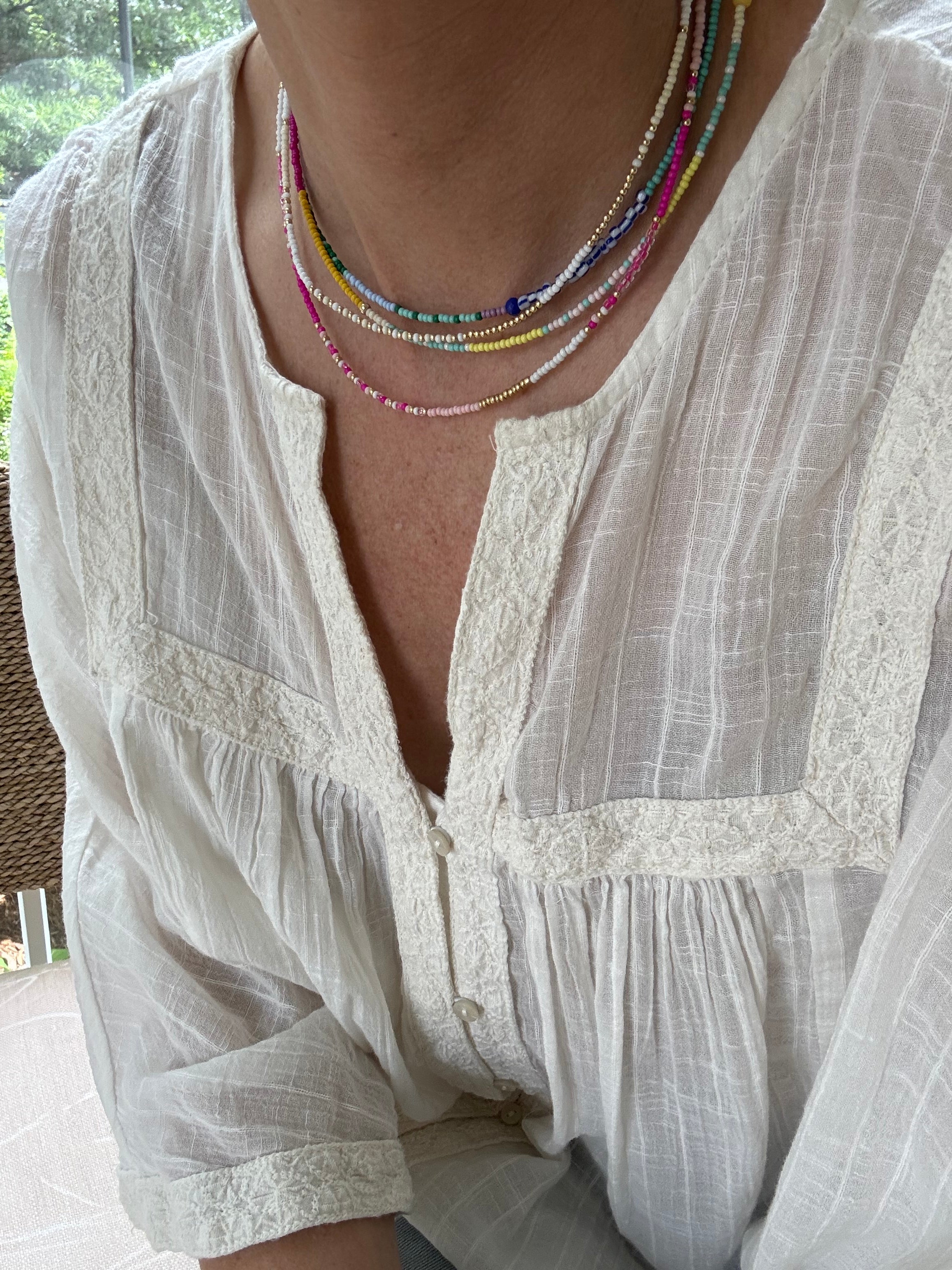 Picture of a woman wearing a white blouse and wearing 4 dainty bead necklaces in colors of white, blues, pinks and golds.