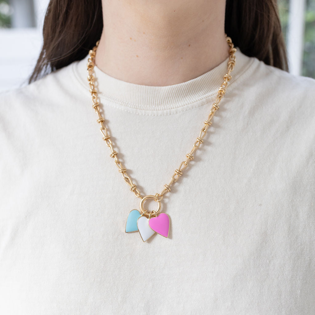 Model wearing gold toggle chain with blue, white and pink heart pendants.