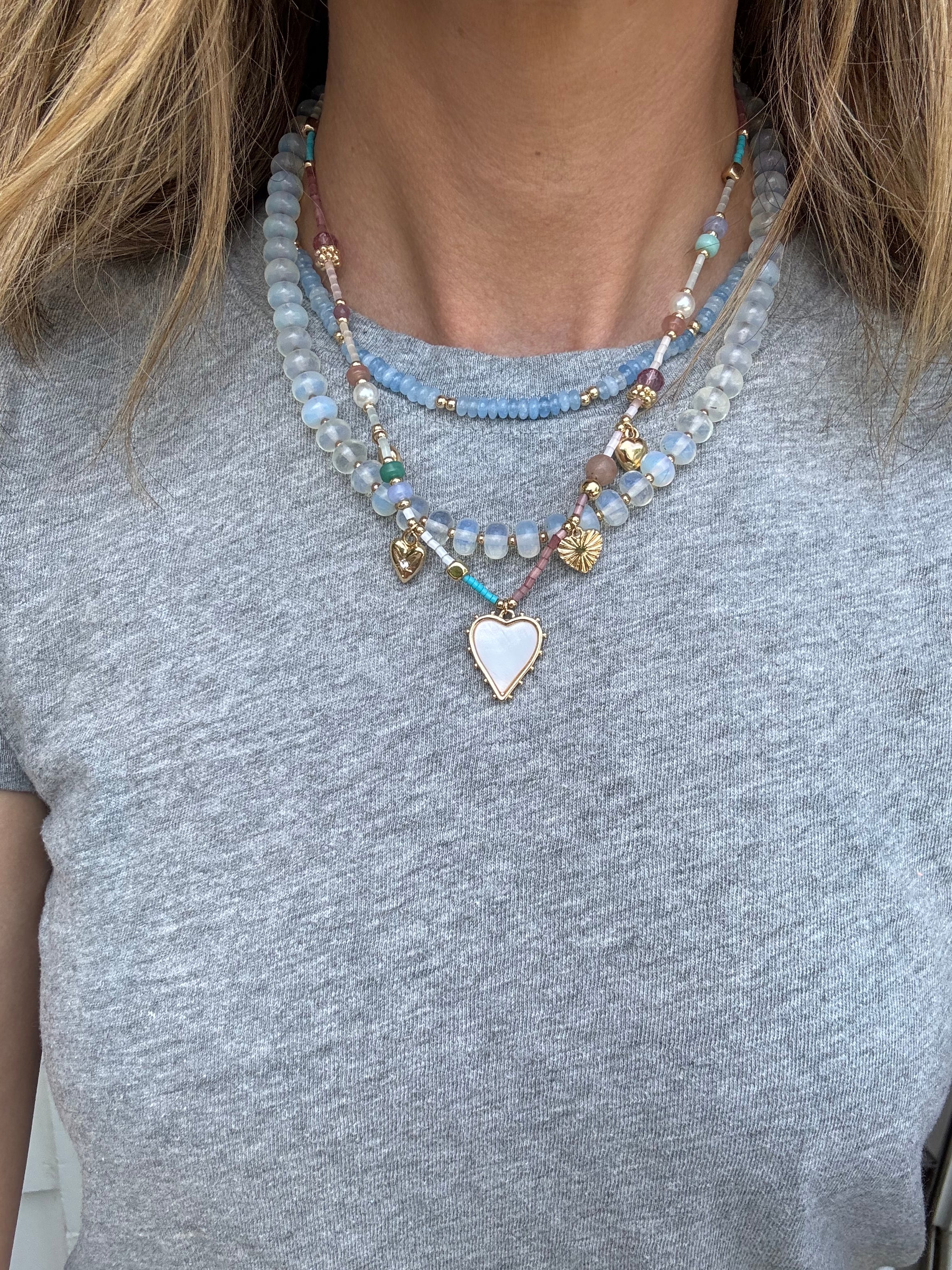 Picture of a woman wearing a gray bead necklace layered with a gold chain with a white enamel heart pendant and a smaller blue bead necklace.