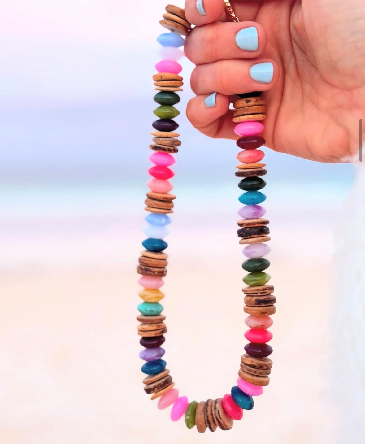 Picture of a hand holding a necklace made of multicolored beads and coconut shell beads. The necklace is made in mostly shades of pinks and blues.