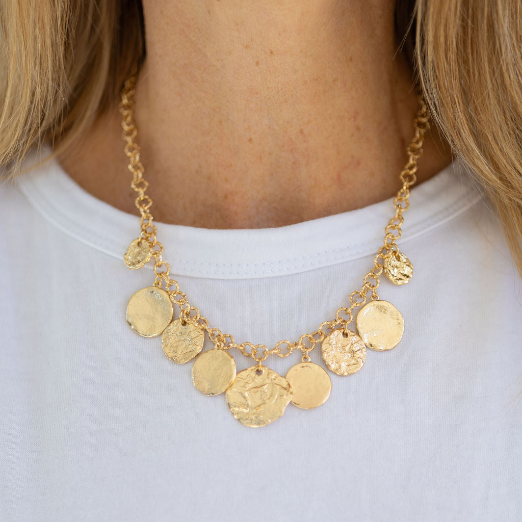 A gold charm necklace with hammered textured discs, displayed on a person wearing a white t-shirt.