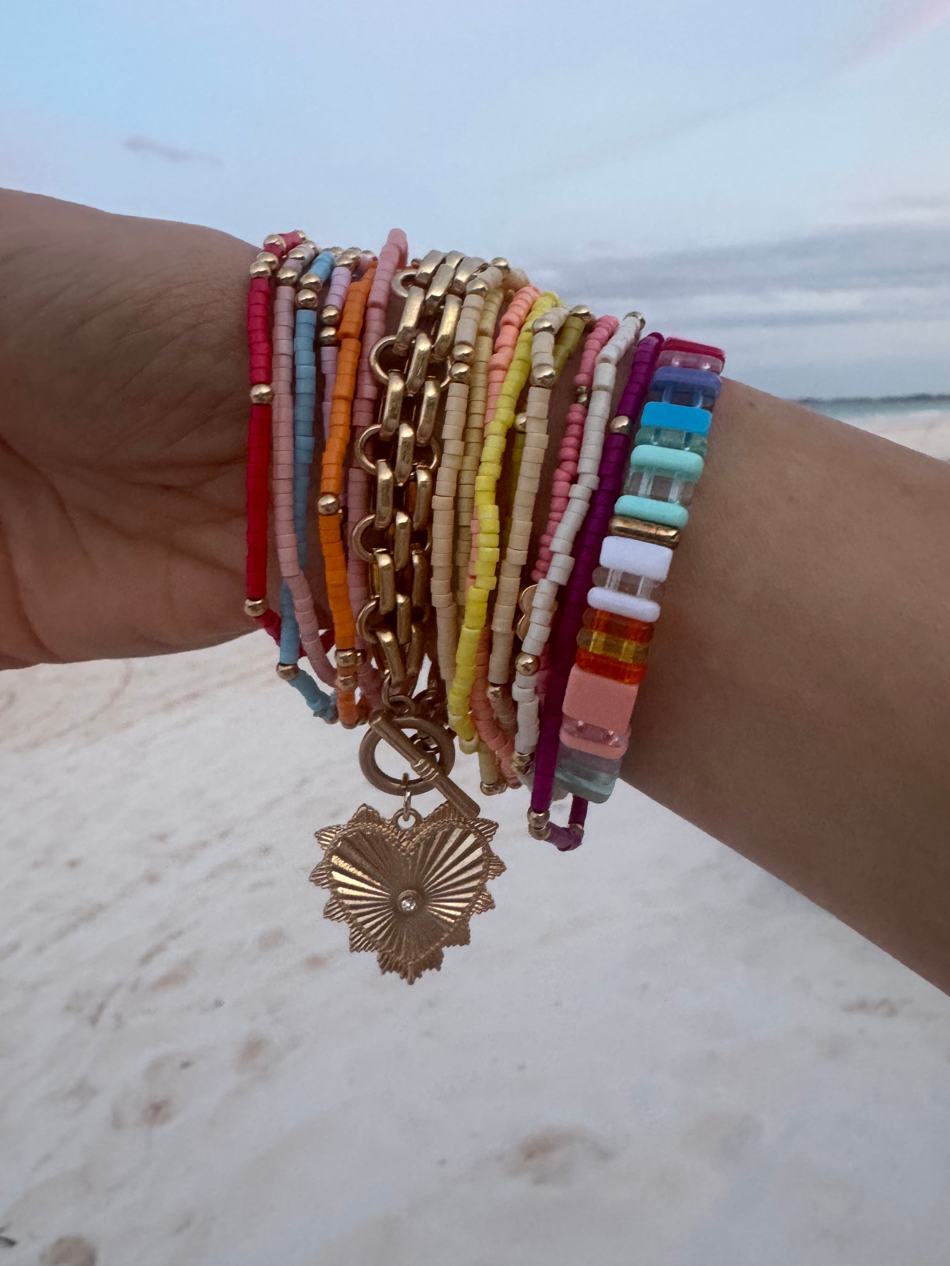 Picture of a woman's wrist wearing 17 different bracelets. 15 bracelets are tiny beaded bracelets of different color with gold accent beads. Bracelets are red, pink, blue, orange, tan, yellow and white. One thick linked gold wrap around bracelet with a large gold heart charm and one multicolored rectangular beaded bracelet.