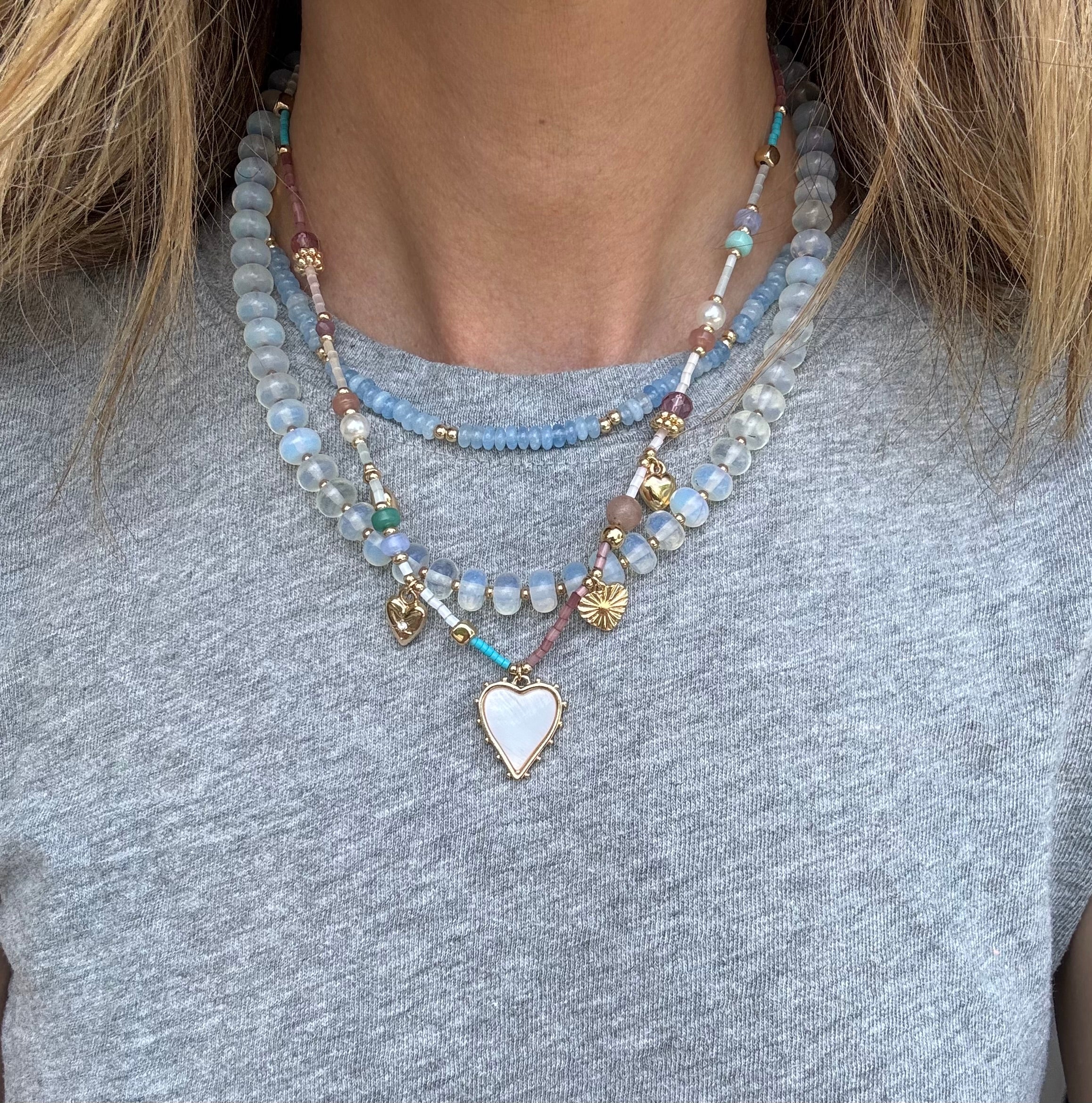 Picture of a woman wearing a gray bead necklace layered with a gold chain with a white enamel heart pendant and a smaller blue bead necklace.