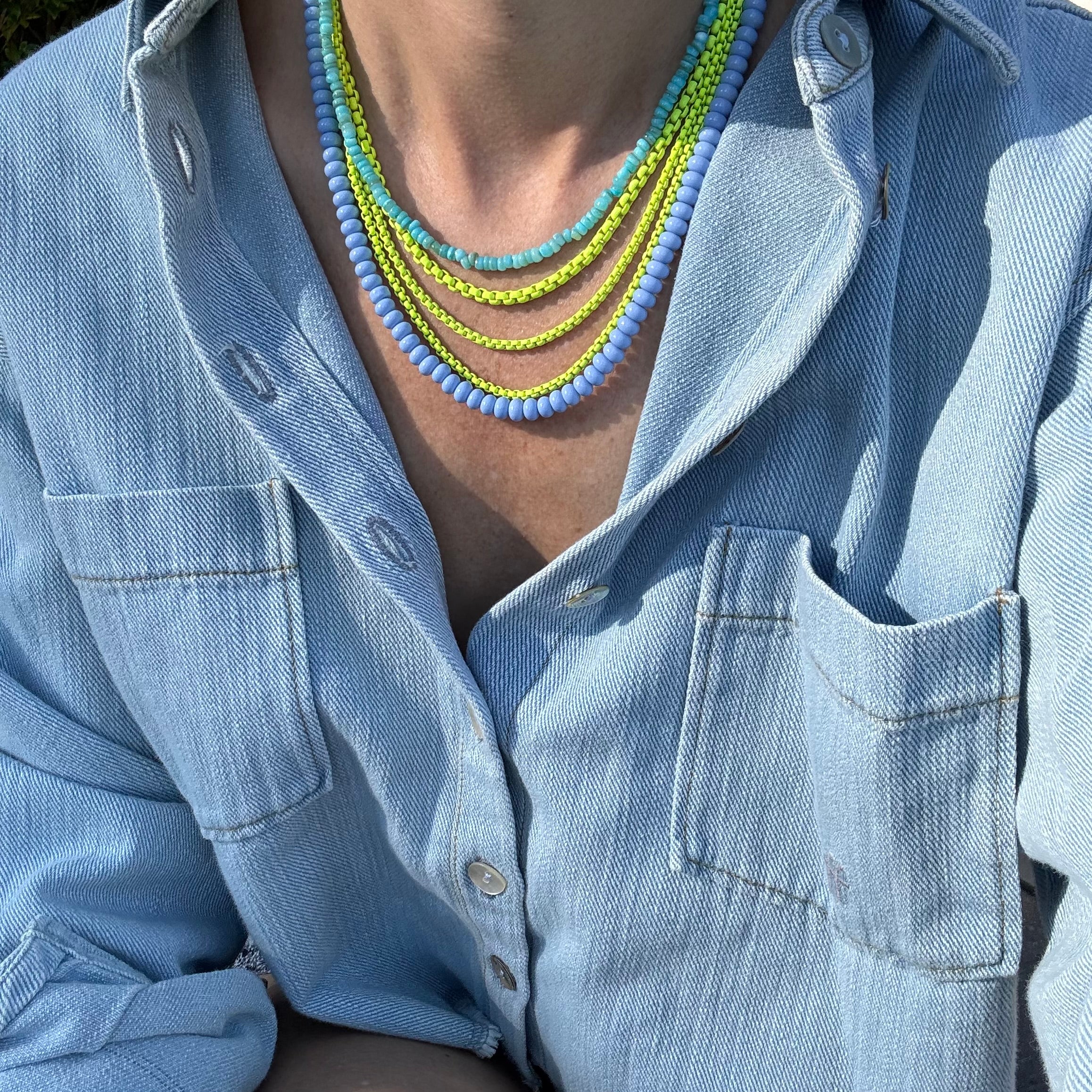 Necklace with blue and yellow beads worn by a person in a light blue denim shirt.