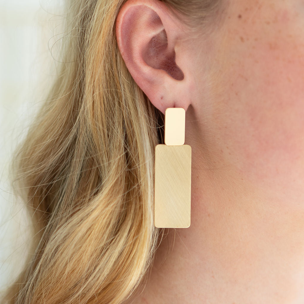 Gold rectangular dangle earring worn by a person with blonde hair.