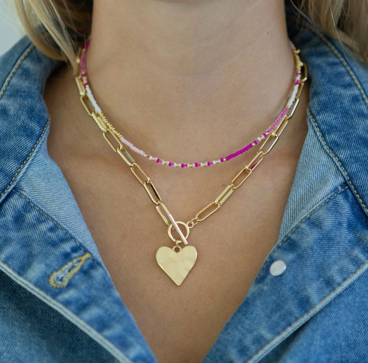 Gold chain link necklace with a gold heart pendant and toggle clasp and a dainty pink and white beaded necklace worn by a person with a denim jacket