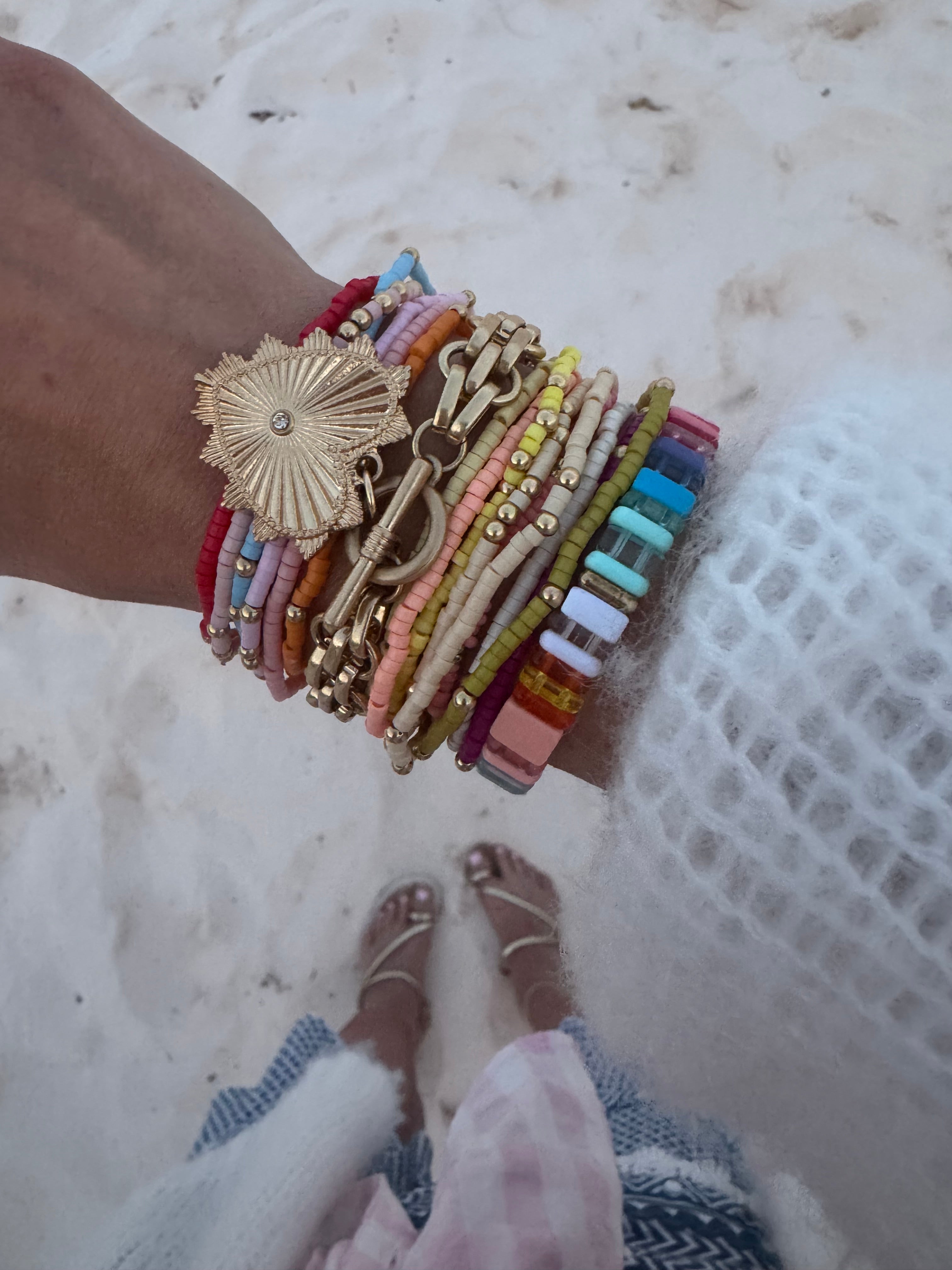 Picture of a woman's wrist wearing 17 different bracelets. 15 bracelets are tiny beaded bracelets of different color with gold accent beads. Bracelets are red, pink, blue, orange, tan, yellow and white. One thick linked gold wrap around bracelet with a large gold heart charm and one multicolored rectangular beaded bracelet.